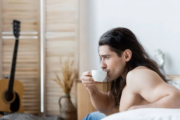 Seitenansicht eines hemdlosen Mannes mit langen Haaren, der Kaffee aus einer Tasse trinkt — Stockfoto