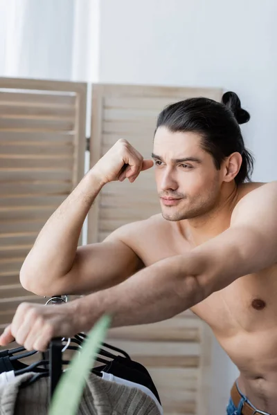 Pensive man with long hair looking away and leaning on rack with hangers — Stock Photo