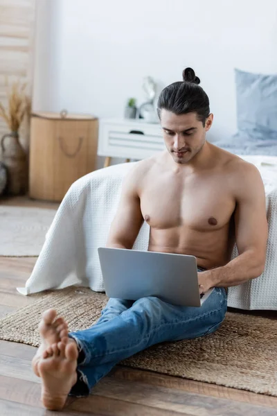 Homme aux cheveux longs assis sur le tapis et utilisant un ordinateur portable près du lit — Photo de stock