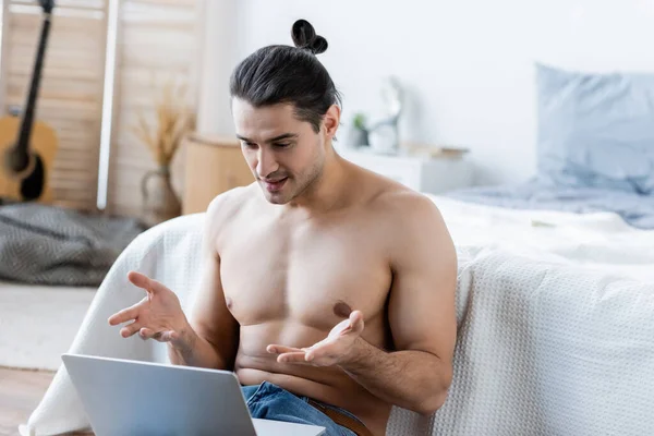 Displeased man with long hair gesturing while using laptop near bed — Stock Photo