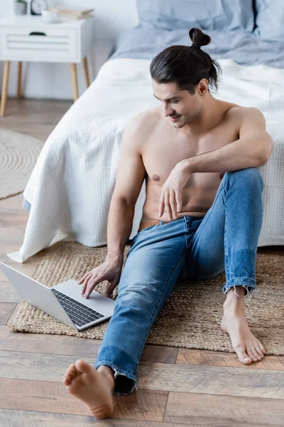 Homem descalço com cabelos longos sorrindo e usando laptop no tapete perto da cama — Fotografia de Stock