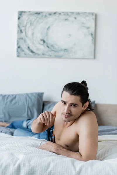 Мужчина без рубашки в синих джинсах лежит на кровати и смотрит в камеру — стоковое фото