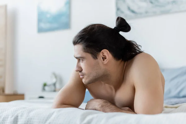 Hombre sin camisa mirando hacia otro lado mientras está acostado en la cama en casa - foto de stock