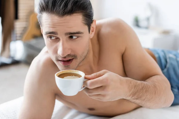 Uomo senza maglietta che tiene in mano una tazza di caffè e riposa sul letto — Foto stock