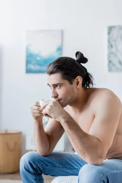 Без рубашки мужчина с длинными волосами держа чашку кофе, сидя на кровати — стоковое фото