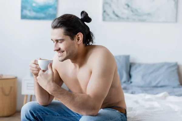 Hombre feliz y sin camisa con el pelo largo sosteniendo taza de café mientras está sentado en la cama - foto de stock