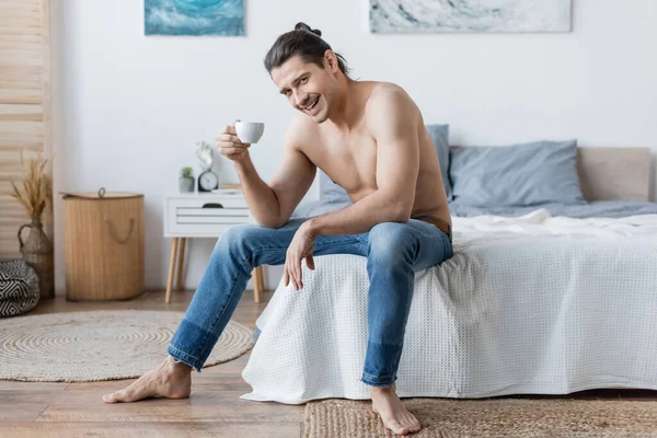 Веселий чоловік з довгим волоссям тримає чашку кави, сидячи на ліжку — стокове фото