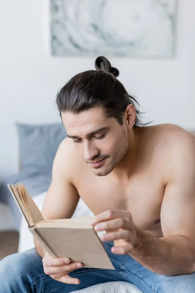 Shirtless man with long hair reading book and sitting on bed — Stock Photo