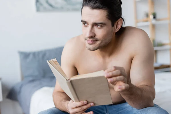 Homem sonhador e sem camisa com livro de cabelo longo segurando no quarto — Fotografia de Stock