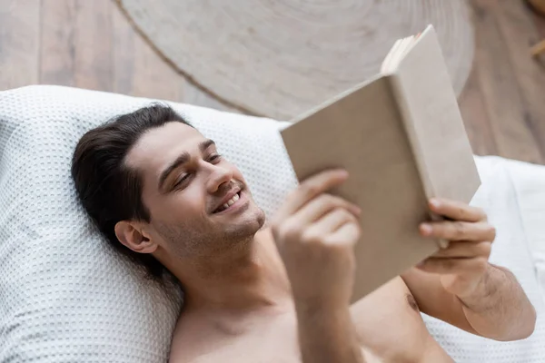Top view of happy and shirtless man reading book while lying on bed — Stock Photo