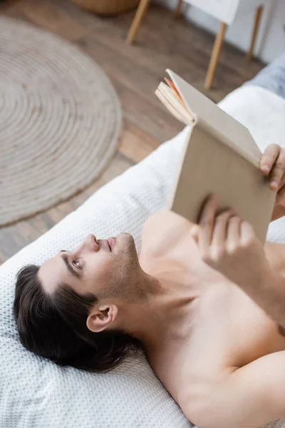 Top view of shirtless man holding book while lying on bed — Stock Photo