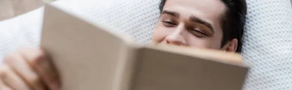 Vista superior do homem cobrindo rosto enquanto livro e deitado na cama, banner — Fotografia de Stock