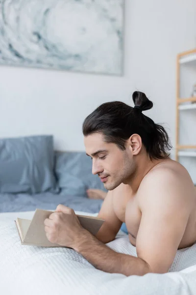 Shirtless man with long hair reading book while resting on bed — Stock Photo