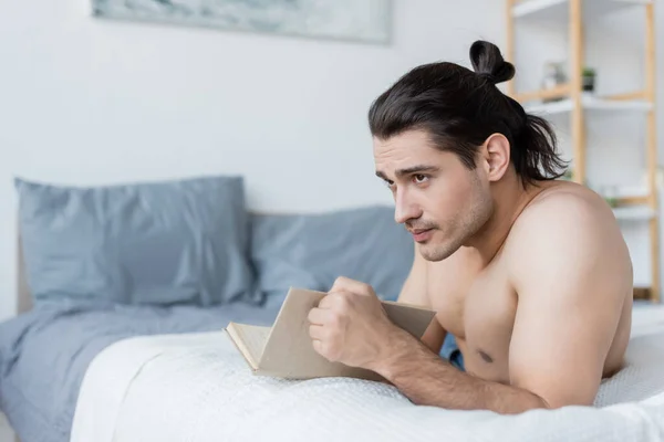 Мужчина без рубашки с длинными волосами, держащий книгу во время отдыха на кровати — стоковое фото