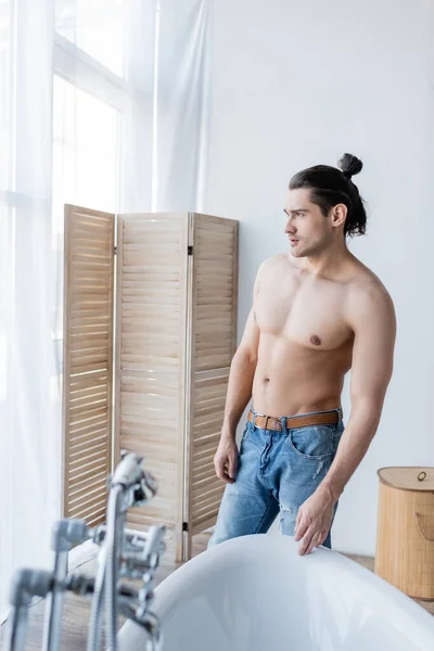 М'язистий чоловік з довгим волоссям, що стоїть в джинсах і дивиться у сучасну ванну кімнату — стокове фото