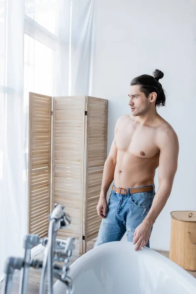 Hombre musculoso con el pelo largo de pie en jeans y mirando hacia otro lado en baño moderno - foto de stock