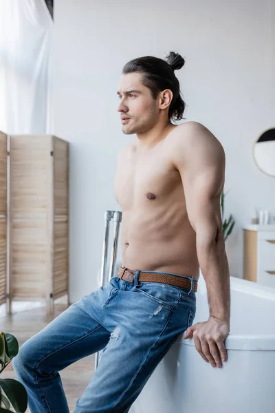 Muskulöser Mann mit langen Haaren sitzt auf Badewanne im modernen Badezimmer — Stockfoto