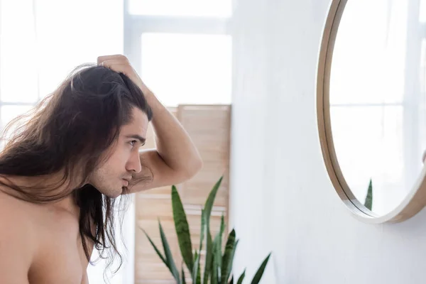 Vista lateral del hombre sin camisa peinado pelo largo cerca del espejo en el baño - foto de stock