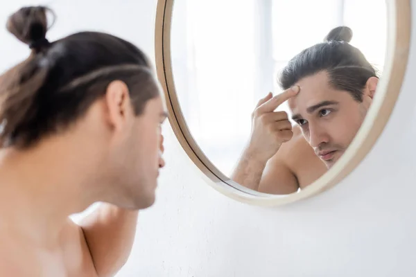 Uomo senza maglietta con i capelli panino sulla testa applicando crema viso e guardando allo specchio — Foto stock