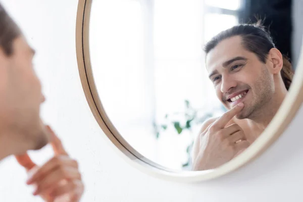Homem feliz e sem camisa sorrindo enquanto olha para o espelho no banheiro — Fotografia de Stock
