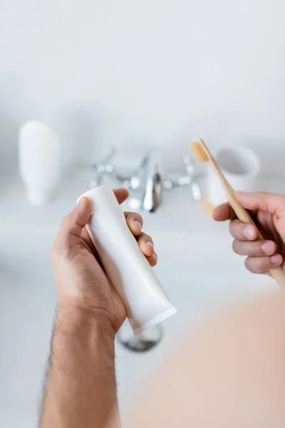 Vista recortada del hombre sosteniendo el tubo de pasta de dientes y el cepillo de dientes en el baño - foto de stock