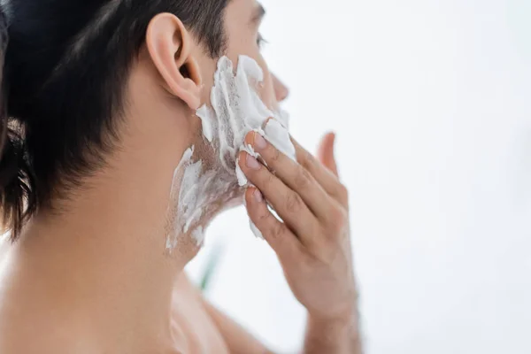 Мужчина без рубашки наносит пену для бритья на лицо в ванной комнате — стоковое фото
