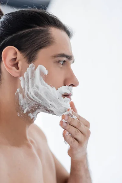 Seitenansicht des hemdslosen Mannes, der im Badezimmer Rasierschaum auf das Gesicht aufträgt — Stockfoto