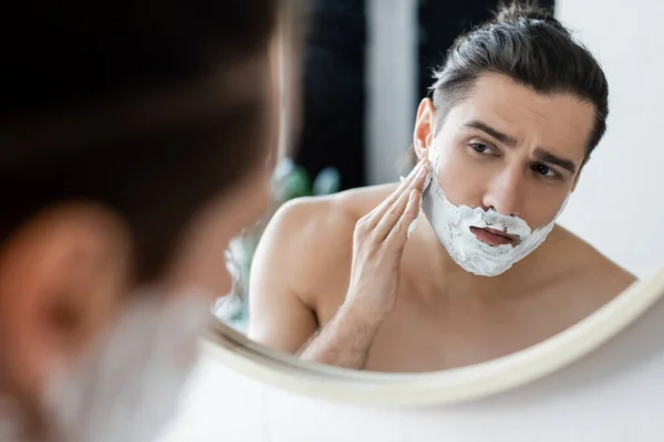 Hemdloser Mann trägt Rasierschaum im Gesicht auf und blickt in den Spiegel — Stockfoto