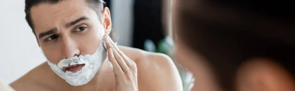 Shirtless man applying shaving foam on face and looking at mirror, banner — Stock Photo