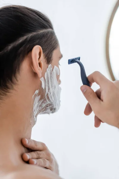 Мужчина без рубашки с пеной для бритья на лице держит безопасную бритву в ванной комнате — стоковое фото