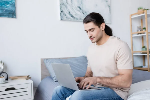 Freelancer with long hair sitting on bed and using laptop — Stock Photo