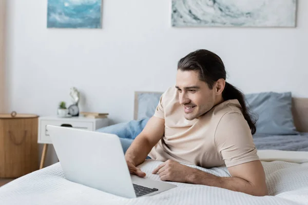 Cheerful freelancer with long hair using laptop in bedroom — Stock Photo