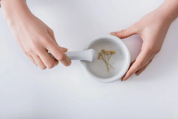 Vista superior de la mujer moliendo flores secas en mortero con pestle en blanco - foto de stock