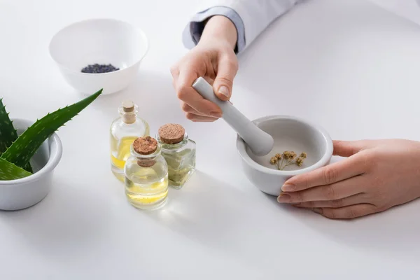 Vista recortada de la mujer moliendo flores secas en mortero con pestle cerca de aloe y botellas - foto de stock