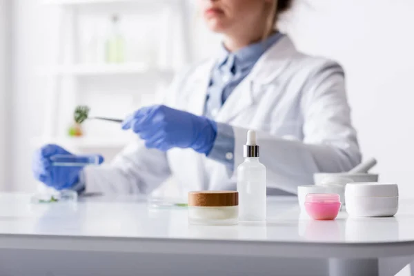 Récipients contenant des produits cosmétiques près de l'assistant de laboratoire flou en gants de latex — Photo de stock
