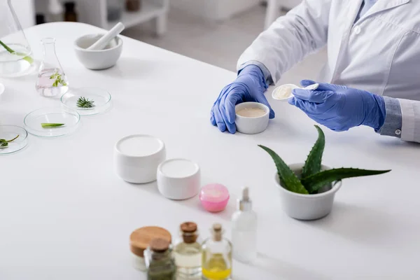 Vista recortada del asistente de laboratorio en guantes de látex sosteniendo cuchara y tazón con crema - foto de stock