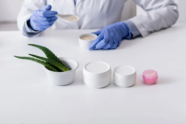 Vista recortada de químico en guantes de látex sosteniendo cuchara y tazón con crema cerca de hojas de aloe y contenedores - foto de stock