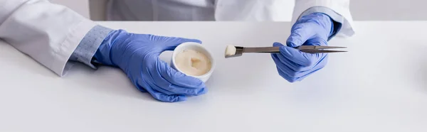 Cropped view of chemist in latex gloves holding tweezers and bowl with cream, banner — Stock Photo