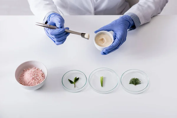 Cropped view of chemist in latex gloves holding tweezers, bowl with cream and fresh plants on test plates near cosmetic powder — Stock Photo