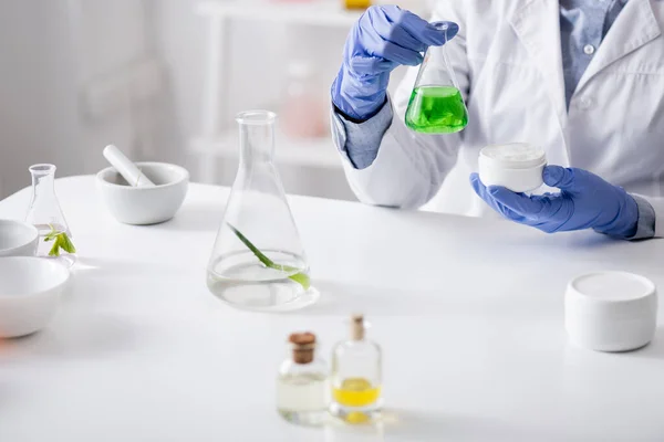 Visão recortada de assistente de laboratório em luvas de látex segurando frasco com líquido verde e creme em recipiente — Fotografia de Stock