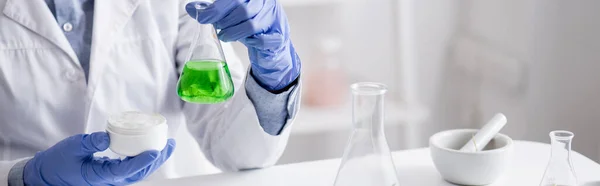 Vista recortada del asistente de laboratorio en guantes de látex sosteniendo matraz con líquido verde y crema en recipiente, banner - foto de stock