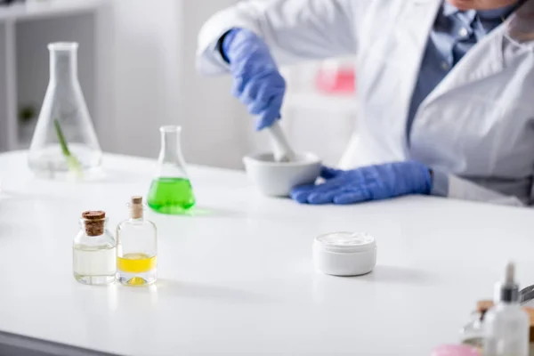 Frascos y botellas cerca de asistente de laboratorio borroso en guantes de látex que trabajan en el laboratorio - foto de stock