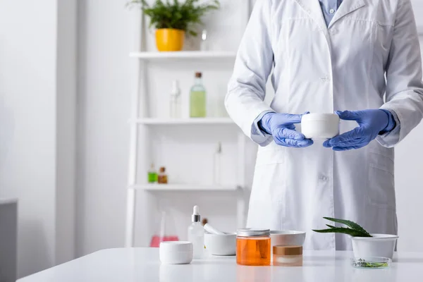 Ausgeschnittene Ansicht einer Laborantin in Latexhandschuhen, die einen Behälter mit Creme in der Nähe kosmetischer Inhaltsstoffe hält — Stockfoto