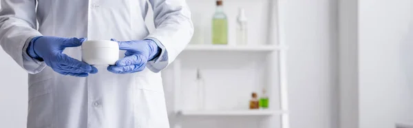 Cropped view of laboratory assistant in latex gloves holding container with cream in lab, banner — Stock Photo