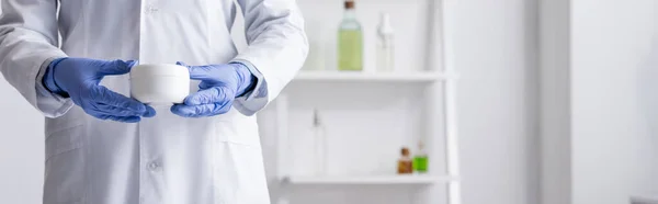 Vue recadrée de l'assistant de laboratoire en gants de latex tenant le récipient avec de la crème en laboratoire, bannière — Photo de stock