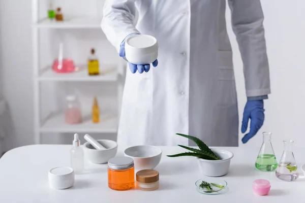 Vista cortada de assistente de laboratório em luvas de látex segurando recipiente com creme perto de ingredientes em laboratório — Fotografia de Stock