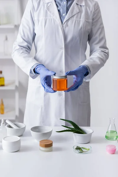 Cropped view of laboratory assistant in latex gloves holding jar with honey near cosmetic products in lab — Stock Photo