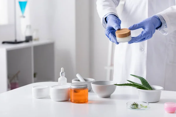 Cropped view of laboratory assistant in latex gloves holding container with cream near cosmetic ingredients in lab — Stock Photo