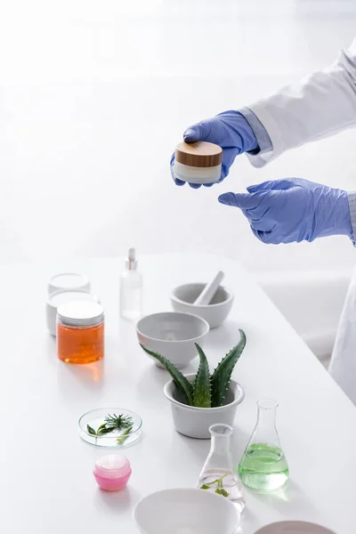 Partial view of laboratory assistant in latex gloves holding container with cream near aloe and cosmetic ingredients in lab — Stock Photo