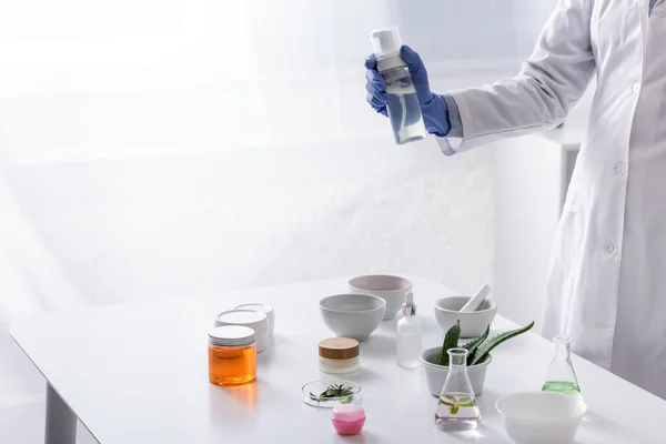 Vista parcial del asistente de laboratorio en guante de látex que sostiene la botella con el producto cosmético cerca de los ingredientes en laboratorio - foto de stock